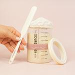 Hand holding a measuring spoon next to a container of powder on a pink background